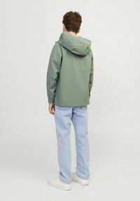 Osoba stojąca tyłem w zielonej kurtce z kapturem, jasnoniebieskich dżinsach i białych trampkach na jednolitym białym tle.