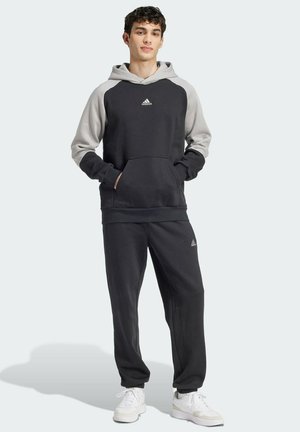 Felpa nera e grigia con cappuccio, dotata di tasca frontale e orlo a coste; abbinata a pantaloni della tuta neri e sneaker bianche.