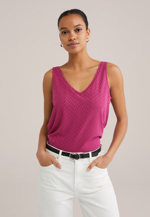 Fuchsia gebreide tanktop met een V-hals en chevronpatroon, gecombineerd met hoge witte broeken en een zwarte riem.