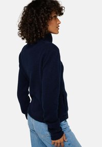 Pull en maille bleu marine avec un col montant, des manches longues et des poignets côtelés, associé à un jean bleu clair. Présente un tissu texturé.