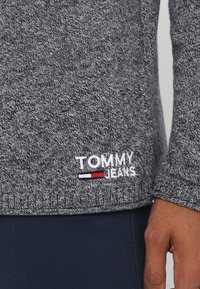 Harmaa neulepaita, jossa on tekstuuri kuviota ja jossa on brodeerattu "TOMMY JEANS" -logo valkoisena, punaisena ja sinisenä helman kohdalla.