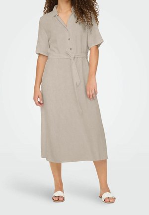 Kobieta w beżowej, zapinanej na guziki sukience midi z krótkimi rękawami i wiązaniem w talii, w połączeniu z białymi, płaskimi sandałami.