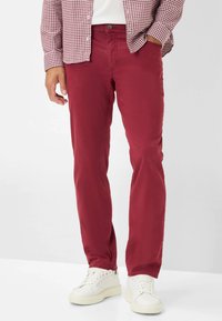 Pantaloni in cotone rossi con vestibilità slim, chiusura con zip e passanti per cintura, abbinati a sneaker bianche. Sfondo bianco semplice.