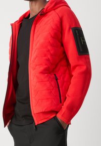 Man med en röd vadderad jacka med dragkedja och en svart ficka på ärmen, svart skjorta och svarta byxor med händerna i fickorna.