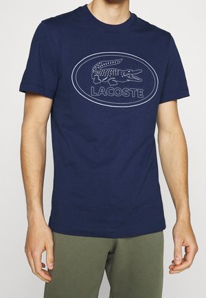 Mann trägt ein marineblaues Lacoste T-Shirt mit weißem Krokodil-Logo und Schriftzug, kombiniert mit olivgrüner Hose.