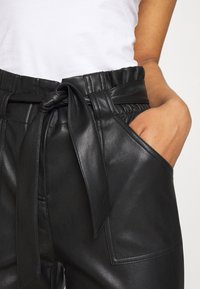 Pantalons en cuir noir taille haute, avec une ceinture à nouer, des poches latérales et une texture lisse. Coutures visibles qui accentuent le tissu.