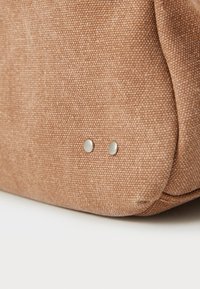 Sac en tissu brun avec une surface texturée et deux clous métalliques sur le côté, mettant en valeur son design durable et ses accents simples.