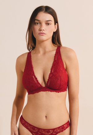 Donna che indossa un reggiseno di pizzo rosso e biancheria intima in pizzo abbinata, in piedi contro uno sfondo beige uniforme.