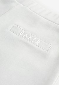 Bílá textilie s texturovaným povrchem, se čtvercovou visačkou s reliéfním nápisem "BAKER" psaným velkými písmeny.