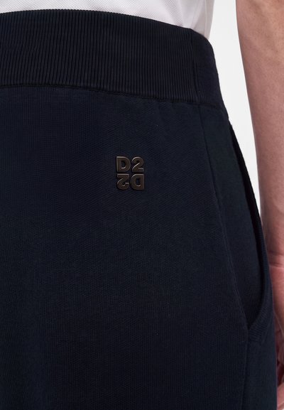 Gros plan d'une personne portant un pantalon bleu marine foncé avec une ceinture côtelée et un logo métallique D2 près de la poche, main reposant à côté de la hanche.