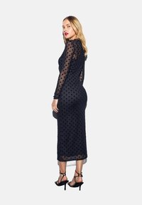 Vestido negro con lunares, de mangas largas y transparentes, silueta ajustada y longitud hasta la mitad de la pantorrilla. Combinado con tacones altos negros con tiras.