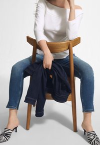 Weiße schulterfreie Bluse, blaue Skinny-Jeans, hält eine dunkelblaue Jeansjacke, schwarz-weiße gestreifte Slipper, sitzt auf einem Holzstuhl.