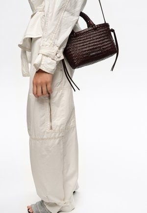 Persona con pantalones y chaqueta sueltos de color blanco roto, llevando un bolso tejido de color marrón oscuro con asas superiores y una correa para el hombro.