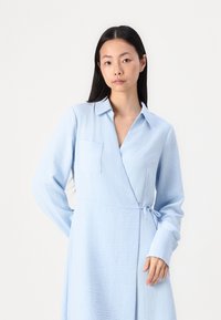 Vestito avvolgente azzurro chiaro realizzato in tessuto testurizzato. Presenta un colletto, maniche lunghe e una tasca frontale con dettaglio in vita a fiocco.
