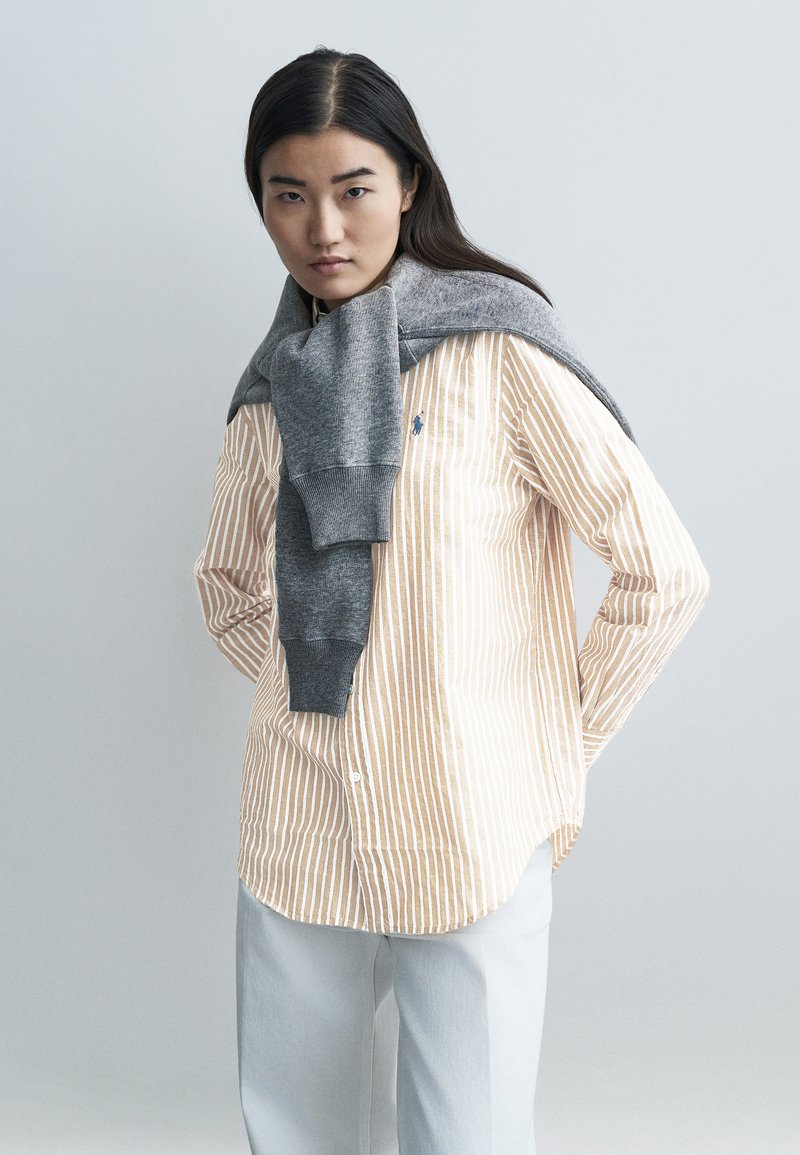 Prúžkovaná béžovo-biela košeľa na gombíky, sivý sveter prehodený cez ramená, kombinovaná s nohavicami svetlosivej farby na neutrálom pozadí.