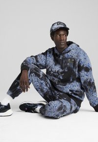 Conjunto de sudadera con capucha y pantalones de chándal con estampado en negro y azul, con un diseño texturizado. Acompañado de una gorra a juego con un logo.