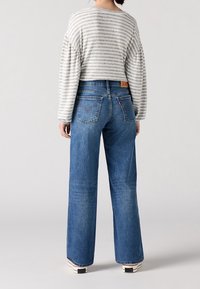 Kvinna som bär blå vida jeans, en långärmad vit och grå randig topp, och vita sneakers, står med ryggen vänd framåt.