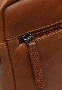 Angolo di una borsa in pelle marrone con chiusura a zip, dotata di un'etichetta piccola con la scritta "The Chesterfield Brand" in rilievo. Superficie liscia e texture.