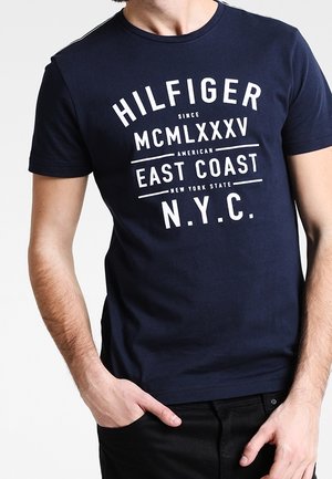 Osoba ubrana w granatowy t-shirt z białym napisem "HILFIGER MCMLXXXV EAST COAST N.Y.C.", stojąca z jedną ręką w kieszeni.