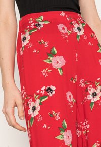 Calças florais vermelhas com uma textura suave, apresentando flores rosas e pretas e folhas verdes contra o vibrante fundo vermelho.