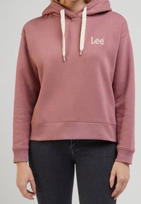 Sweat à capuche rose pâle avec cordons blancs et logo "Lee" brodé. Présente une coupe décontractée, des poignets côtelés et une texture de tissu lisse.