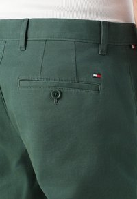 Zelené bavlnené nohavice so zadnou vreckom, ktoré sa zapína na gombík a majú malú nášivku s logom v červenej, bielej a modrej farbe blízko vrecka.