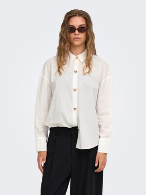 Femme aux cheveux ondulés portant des lunettes de soleil foncées, une chemise écrue à boutons en forme de cœur et un pantalon noir à jambes larges.