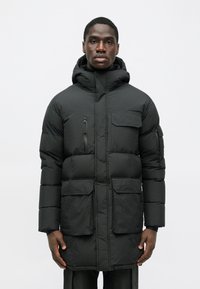 Črna puhovka z kapuco, ki ima dve veliki sprednji žepki, majhen zadrževalni žep na zgornji levi strani in prešito teksturo.