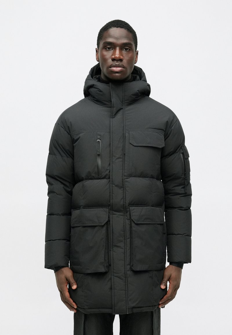 Črna puhovka z kapuco, ki ima dve veliki sprednji žepki, majhen zadrževalni žep na zgornji levi strani in prešito teksturo.