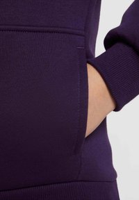 Sudadera morada con una mano en un bolsillo frontal. Presenta puños de canalé y una tela suave y texturizada. Diseño simple y funcional.