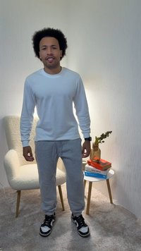 Man met een wit shirt met lange mouwen en grijze joggingbroek, staand op een tapijt nabij een stoel en een bijzettafel met boeken en een vaas.