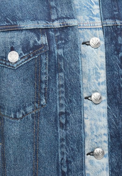 Casaco de ganga com vários tons de azul, botões prateados, um bolso no peito e um acabamento ligeiramente texturizado com costuras visíveis.