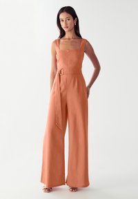 Žena v bezrukávovém terakotovém kombinézovém modelu s širokými nohavicemi, čtvercovým výstřihem a páskem ze stejného materiálu s kovovým kroužkem.