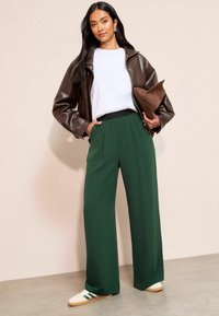 Donkergroene wijde broek met een zwarte elastische tailleband, gecombineerd met een wit T-shirt en een donkerbruine leren jas, terwijl een bruine clutch wordt vastgehouden.