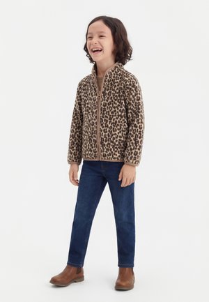 Dieťa sa smeje, stojí a má na sebe bundu so zipsom s leopardím vzorom, modré džínsy a hnedé členkové topánky na jednoduchom bielom pozadí.