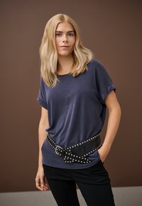 Fiatal nő szőke hajjal, laza sötétkék ingben, szegecselt fekete övvel és fekete nadrágban, barnás háttér előtt áll.