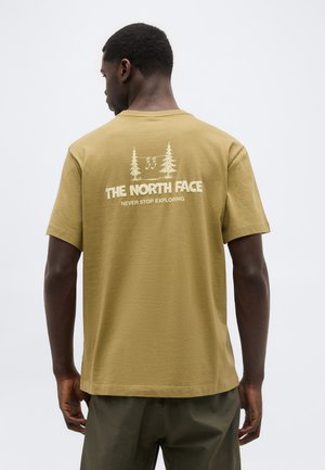 Hombre vistiendo una camiseta amarillo mostaza con el texto "THE NORTH FACE" y un diseño de árbol, visto desde atrás sobre un fondo claro y liso.