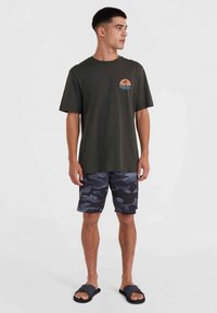 Groene katoenen T-shirt met een klein gedrukt logo, gecombineerd met zwarte camouflage zwembroeken. Het model draagt donkere slippers. Eenvoudig ontwerp en pasvorm.
