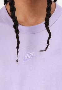 Levandų spalvos megztinis su apvalia iškirpte, su mažuoju baltu siuvinėtų Nike logotipu. Aplink kaklą yra laisvos pynės. Minkšta medvilnės tekstūra.