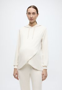 Φόρμα εγκυμοσύνης σε ανοιχτό μπεζ χρώμα με σχεδιασμό τύπου wrap, μακριά μανίκια και κουκούλα με κορδόνι. Μαλακό ύφασμα με ομαλή υφή. Συνοδεύεται από παντελόνι που ταιριάζει.