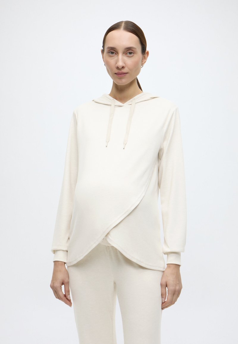 Φόρμα εγκυμοσύνης σε ανοιχτό μπεζ χρώμα με σχεδιασμό τύπου wrap, μακριά μανίκια και κουκούλα με κορδόνι. Μαλακό ύφασμα με ομαλή υφή. Συνοδεύεται από παντελόνι που ταιριάζει.