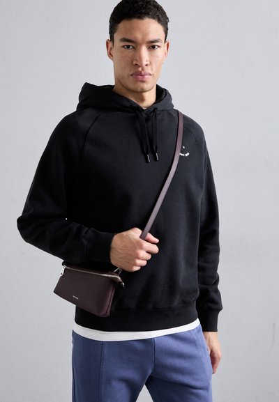 Sweatshirt preto com capuz e cordões, usado sobre uma camisa branca. O modelo segura uma pequena bolsa de ombro retangular em bordeaux com fecho.