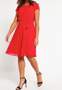 Une robe rouge, à mi-mollet, en tissu léger avec un corsage ajusté, des manches courtes et une ceinture à nouer. Associée à des talons noirs à brides aux chevilles.