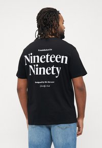 Zwarte katoenen t-shirt met achterop de tekst: "Opgericht in negentien negentig," en "Ontworpen door de J&J crew" in het wit. Korte mouwen, normale pasvorm.