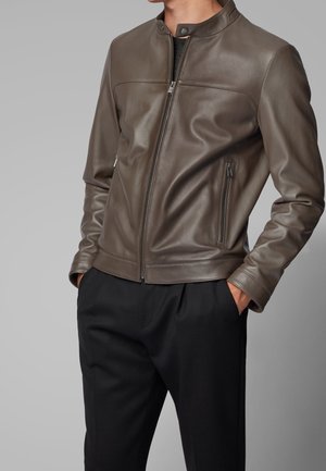 Mann trägt eine braune Lederjacke mit Reißverschluss und Reißverschlusstaschen an den Seiten sowie schwarze Hosen, Hände in den Taschen, grauer Hintergrund.