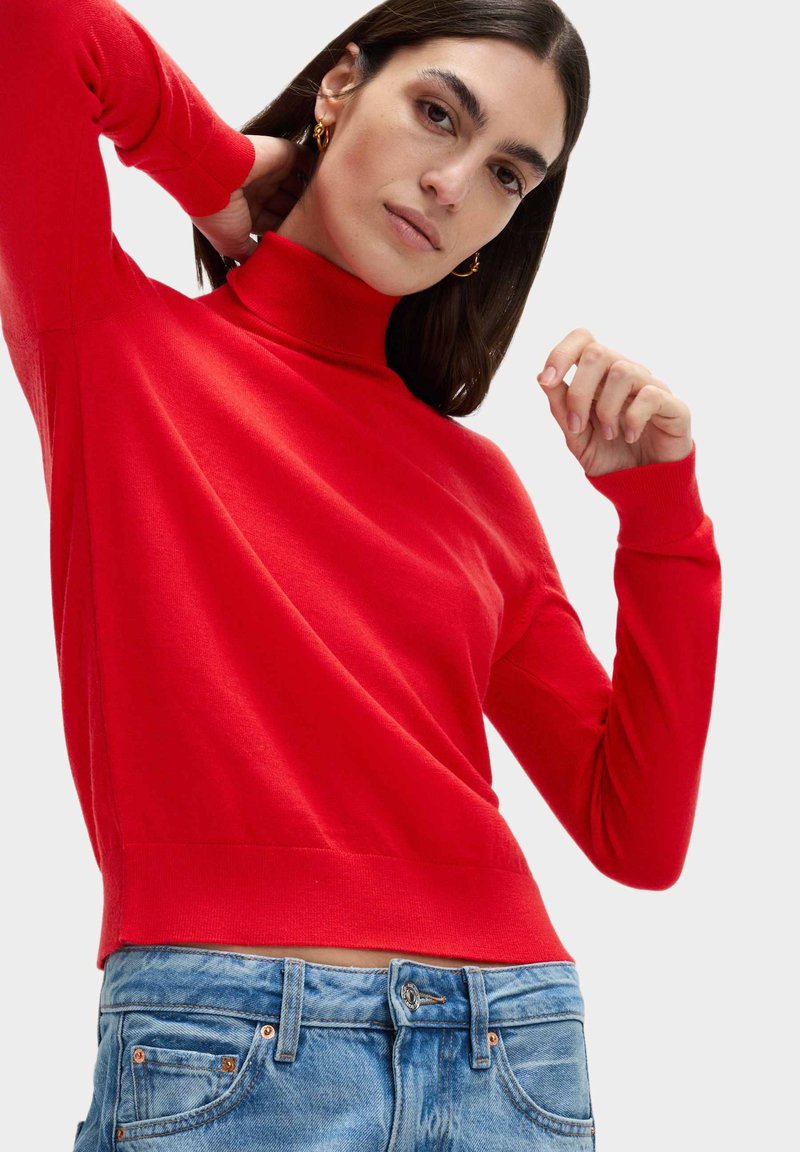 Maglione rosso a collo alto realizzato in materiale morbido, con maniche lunghe, polsini a costine e una vestibilità aderente. Abbinato a jeans in denim blu chiaro.
