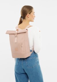 Zaino roll-top in morbido tessuto rosa, dotato di spallacci regolabili, una tasca frontale con zip e accessori laterali per una chiusura sicura.