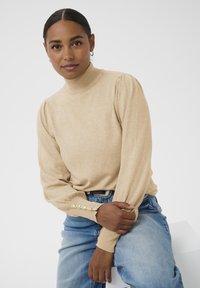 Tumehise juustega naine, kes kannab beeži kõrge kaelusega kampsunit kuldsete nööpidega ja helesiniseid teksaseid, istub käsi oma käsivarrel.