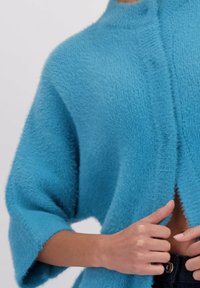 Pull turquoise surdimensionné avec des manches courtes, fabriqué en matériau doux et duveteux. Design à l'avant ouvert, finition texturée et forme décontractée.