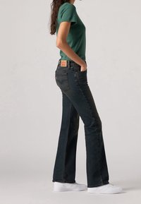 T-shirt vert à manches courtes et jean évasé bleu foncé. Le tissu du t-shirt semble lisse ; le jean présente un design classique à cinq poches avec un patch en cuir.